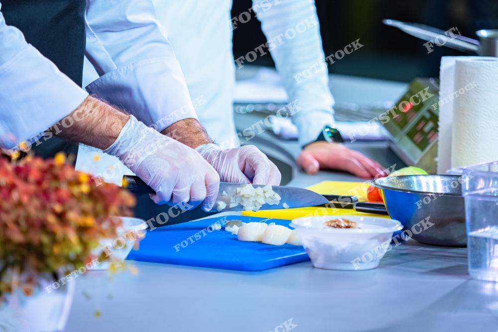 Профессиональный повар в белой униформе и перчатках нарубает лук на синей разделочной доске в кухне ресторана. Рядом стоят тарелки с другими ингредиентами и блюдами, а также зелень, добавляющая яркости интерьеру. Видны кухонные принадлежности и оборудование, в том числе ножи и миски.